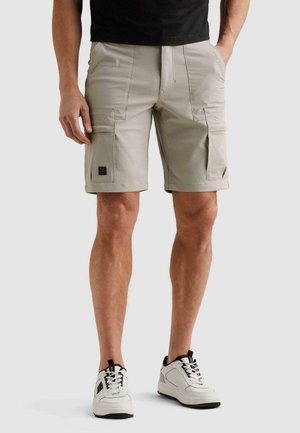 Man draagt beige cargoshort met zakken en witte sneakers, staand tegen een effen lichte achtergrond.