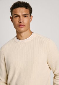 Kremna rebrasta majica z okroglim izrezom in teksturirano tkanino. Obuta čez belo srajco, ima oprijet kroj in vidno pleteno zasnovo.