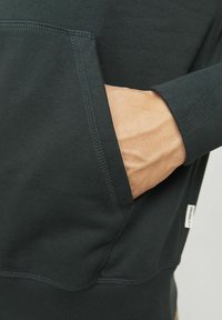 Ciemnozielona bluza z kieszenią kangurką z przodu, ściągaczami przy rękawach oraz widoczną metką po dolnej stronie. Miękka, gładka struktura.