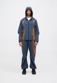Waterdicht jack en broek set in marineblauw met bruine accenten, met rits aan de voorkant, capuchon en twee zijzakken. Textuur afgewerkt stof.