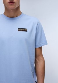 Lys blå bomulds T-shirt med korte ærmer, med et rektangulært sort logo-patch med hvid og rød tekst på venstre brystområde.