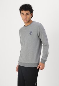 Hanorac gri cu decolteu rotund, manșete și tiv cu grătar. Prezintă o etichetă logo bleumarin pe pieptul stâng. Fabricat dintr-un material moale, texturat.