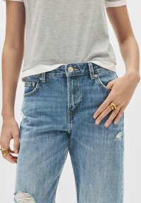 Jeans in denim azzurro chiaro con effetti usurati e vita alta, caratterizzati da una chiusura con bottone, un design a cinque tasche e leggero sbiadimento. Indossati con una maglietta grigia.