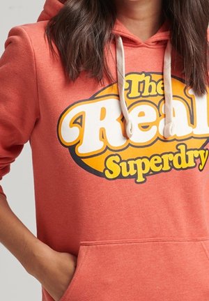 Personne portant un sweat à capuche corail avec des cordons blancs et un grand motif jaune et blanc indiquant "The Real Superdry" à l'avant.