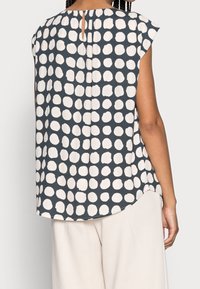 Blusa a maniche corte di colore blu scuro con pois color crema. Presenta un'apertura a chiave sul retro e un orlo curvo. Tessuto morbido al tatto.