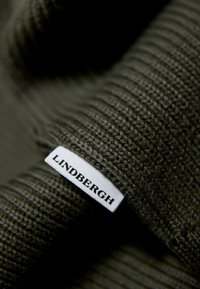 Olivgrüner Strickstoff mit einem strukturierten Muster, versehen mit einem weißen Etikett, auf dem der Text "LINDBERGH" in Schwarz steht.