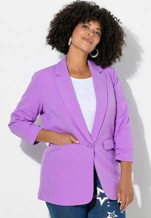 Laventelinvärinen bleiseri, jossa yksittäinen nappi, lovikaulus ja kolmen neljänneksen hihat, yhdistettynä valkoiseen toppiin ja tähtikoristeellisiin sinisiin farkkuihin.