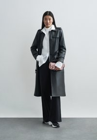 Svart lær trenchcoat med knappelukking, over en hvit skjorte og svarte vide bukser. Holder en liten rosa håndveske. Hvit sneakers.