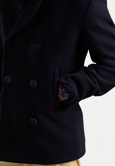 Manteau en laine bleu marine avec fermeture à double boutonnage, poches ajustées et une montre visible dans la poche principale. Texture lisse.