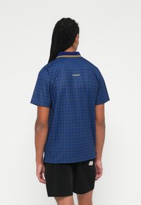 Kortærmet polo-shirt med blåt ternet mønster, gule kraveapplikationer og "PROJEKTER" broderet på ryggen. Bomuldsstof.