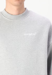 Grey sweatshirt made of cotton blend with ribbed collar. Features small white embroidered brand logo on the left chest. Textured finish.