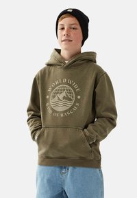 Olivgrön hoodie med en framficka, prydd med en cirkulär grafisk design med text. Matchas med ljusblå denimbyxor.