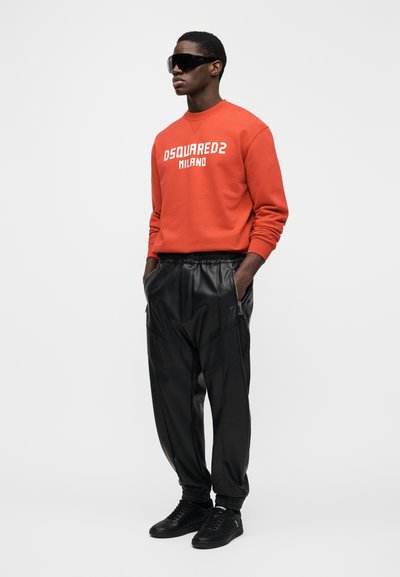 Oranžová mikina s bílým nápisem "DSQUARED2 MILANO"; černé joggery z umělé kůže; černé tenisky; model nosí tmavé velké sluneční brýle.