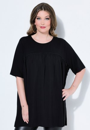 Olkapäihin ulottuvat hiukset omaava nainen, joka käyttää väljää mustaa lyhythihaista mekkoa ja suuria rengaskorvakoruja, poseeraa yhdellä kädellä lantiolla.