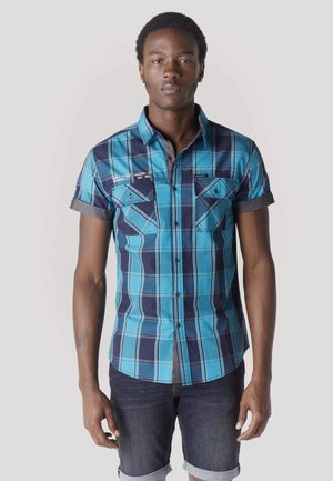 Giovane uomo che indossa una camicia a quadri blu a maniche corte con bottoni e pantaloncini di jeans scuri arrotolati, in piedi contro uno sfondo semplice.