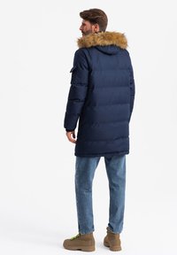 Giacca imbottita blu navy con cappuccio foderato di pelliccia, dotata di zip frontale e tasche laterali, indossata sopra jeans di denim chiaro e stivali marroni.
