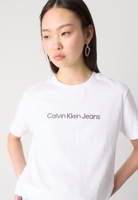 Balta medvilninė marškinėliai su tamsiu atspaudu "Calvin Klein Jeans", apvaliu kaklu ir trumpomis rankovėmis, pristatyti neutralioje fone.