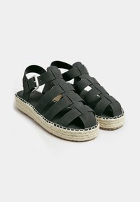 Sandali espadrillas neri con tomaia intrecciata, cinturino regolabile alla caviglia e suola intermedia in juta. Presentano una superficie testurizzata e cuciture a contrasto.