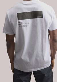 Bílá bavlněná tričko s černým obdélníkovým motivem na zadní straně s textem, krátké rukávy a kulatý výstřih. Minimalistický design.