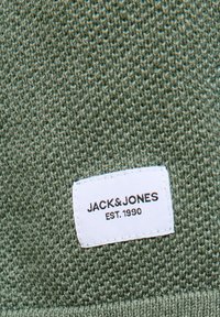Green knitted fabric with a white rectangular label stitched on, displaying "JACK&JONES EST. 1990" in black text.
