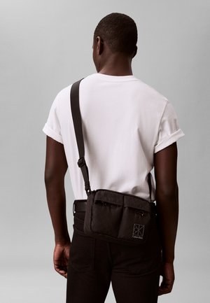 Persoon draagt een wit T-shirt en een zwarte broek, met een zwarte crossbodytas van Calvin Klein met een verstelbare band over de rug.