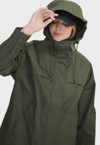 Veste imperméable vert olive avec une capuche, zip avant et cordons réglables. Comprend de grandes poches poitrines et des poignets élastiques.