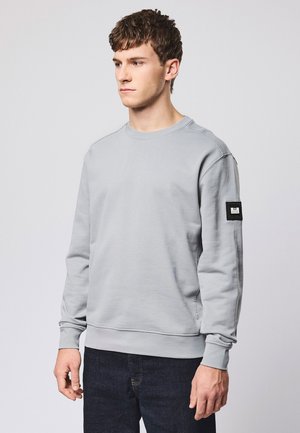 Młody mężczyzna w jasnoszarej bluzie z czarną naszywką na lewym rękawie, ubrany w ciemnoniebieskie dżinsy, stoi na tle jednolitego tła.