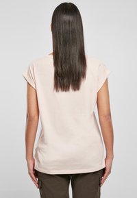 Osoba z długimi prostymi ciemnymi włosami, ubrana w jasnoróżową koszulę z krótkim rękawem i ciemne spodnie cargo, stojąca tyłem na jednolitym tle.