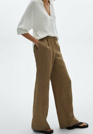 Frau trägt lockere braune Nadelstreifenhose, weiße Bluse mit hochgekrempelten Ärmeln und schwarze Sandalen, steht mit den Händen in den Taschen.