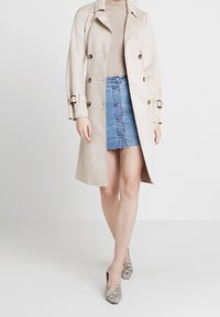 Trench-coat beige en tissu doux avec des boutons croisés, accompagné d'une jupe en denim et de chaussures à imprimé serpent.