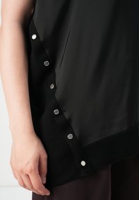 Svart topp med en silkeaktig glans, med ribbestrikket kant og metallisk trykknappåpning langs den ene siden, som fremhever en raffinert design.
