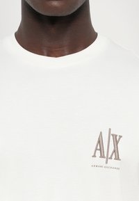 Fehér pamut póló kerek nyakkivágással és rövid ujjakkal, a mellkason egy barna hímzett "A/X" és "ARMANI EXCHANGE" logóval.