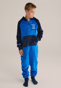 Pojke iklädd en blå och marinblå fleeceoverall med en "R"-applikation, stående barfota, tittande åt vänster, med händerna i fickorna.