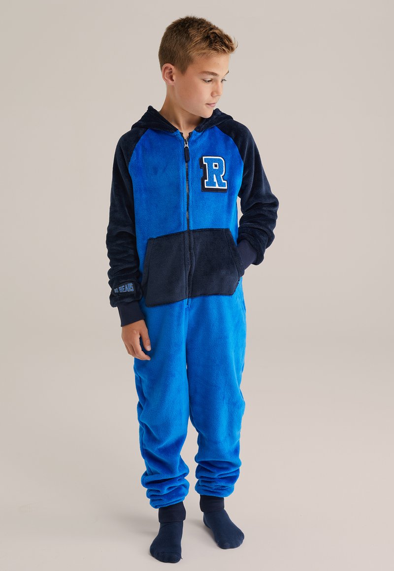 Pojke iklädd en blå och marinblå fleeceoverall med en "R"-applikation, stående barfota, tittande åt vänster, med händerna i fickorna.