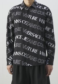 Svart långärmad skjorta med stora vita textmönster i varierande typsnitt. Tyg verkar vara lättviktigt med en smidig textur.