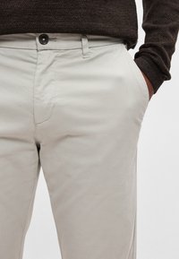 Heledad hallid puuvillased püksid sirge lõikega, millel on nupuga kinnis, küljetaskud ja sile tekstuur.