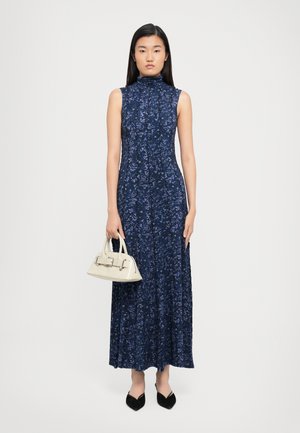 Navy floral sleeveless maxi dress with a high collar, pleated texture, complemented by a small cream handbag and pointed black flats.