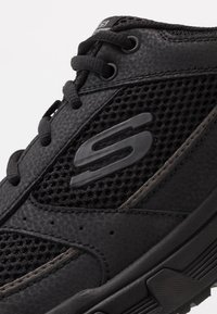 Zapatilla deportiva negra con material de malla y sintético, acabado texturizado, forma redondeada y un logo prominente en el lateral.