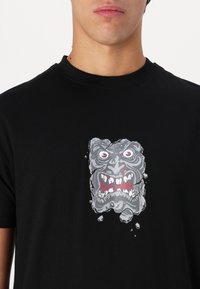 Zwarte t-shirt met korte mouwen met een grafisch ontwerp van een woest gezicht, voornamelijk grijs met rode accenten, tegen de zwarte stof.