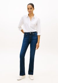 Mulher vestindo uma camisa branca de botões com as mangas dobradas, jeans azul escuro e tênis brancos, em pé com uma mão atrás das costas.