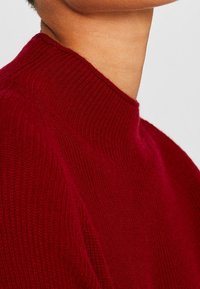 Gros plan sur une personne portant un pull rouge côtelé à col haut, montrant l'épaule, le cou et une partie de la mâchoire.