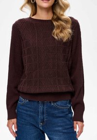 Brązowy sweter z dzianiny o teksturowanym wzorze w kształcie V, z prążkowanymi mankietami i dopasowanym stanem. Połączony z niebieskimi dżinsami.