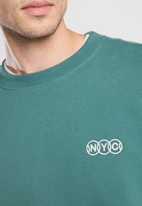 Teal sweatshirt featuring a round neckline and embroidered "NYC" logo in white. Smooth texture and casual design.