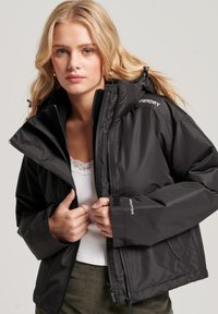 Zwarte geïsoleerde jas met een hoge kraag, rits aan de voorkant, verstelbare manchetten en logo-detail op de schouder en mouw.