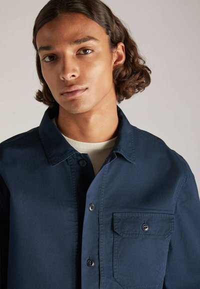 Un giovane con capelli ondulati appare, indossando una camicia button-up blu scuro con taschino sul petto, su uno sfondo grigio morbido.