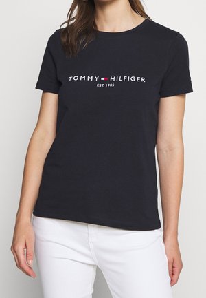 Femme portant un t-shirt noir à manches courtes Tommy Hilfiger avec le logo et le texte "EST. 1985", associé à un pantalon blanc.