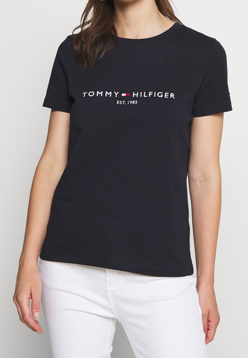 Žena v černém tričku s krátkým rukávem od Tommy Hilfiger se znakem a textem "EST. 1985", v kombinaci s bílými kalhotami.