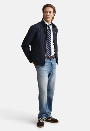 Quilted navy jacket over a white shirt and striped tie. Light blue jeans and brown sneakers with white accents. Casual fit and textured materials.