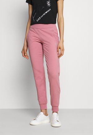Femme portant un pantalon de jogging rose, des baskets blanches et un t-shirt noir avec un texte graphique, debout devant un mur clair uni.