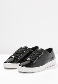 Baskets basses en cuir verni noir avec lacets noirs, œillets dorés, détail du logo doré et semelles en caoutchouc blanches sur une surface blanche.